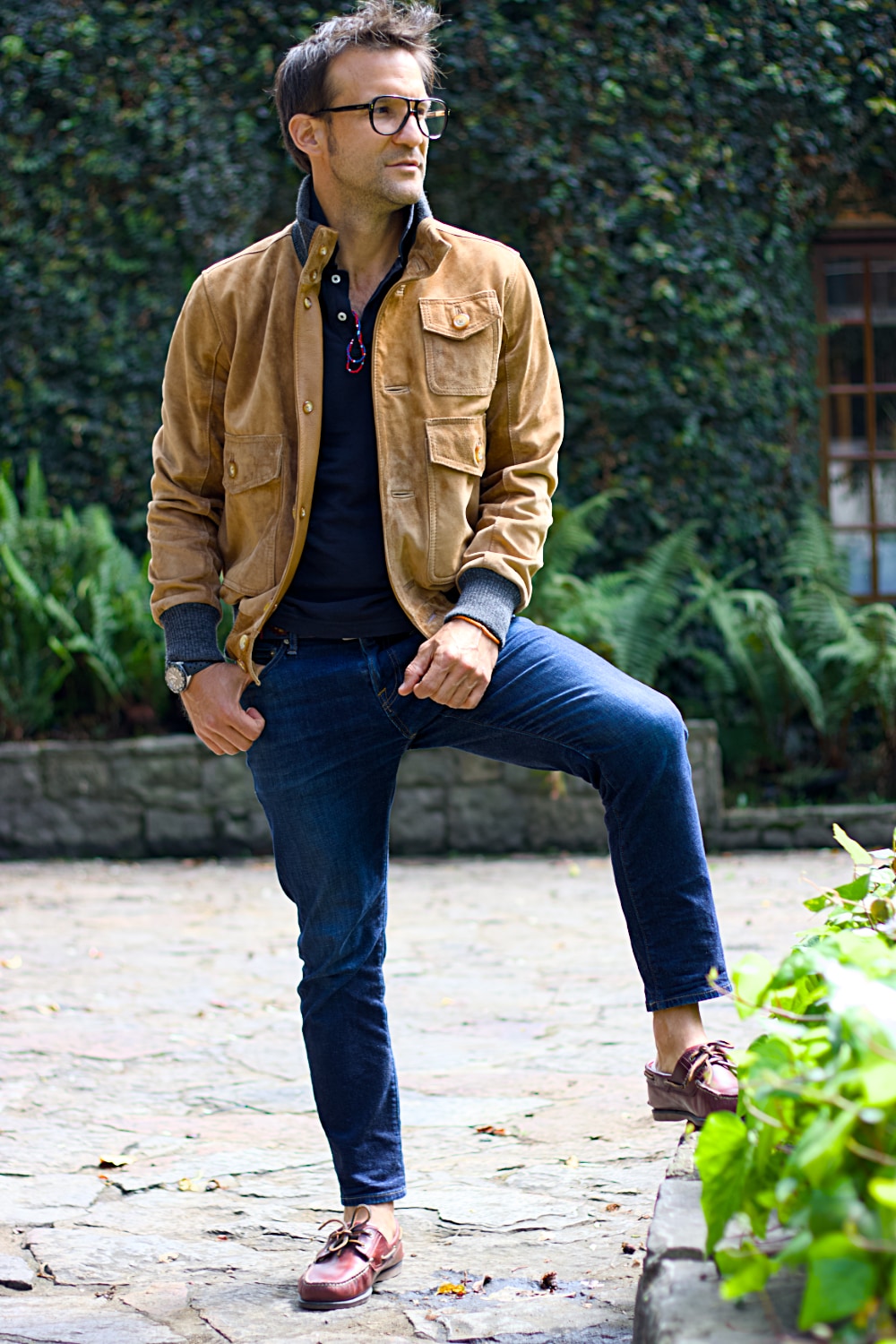 Man wearing a brown jacket and blue jeans standing outdoors with greenery in the background